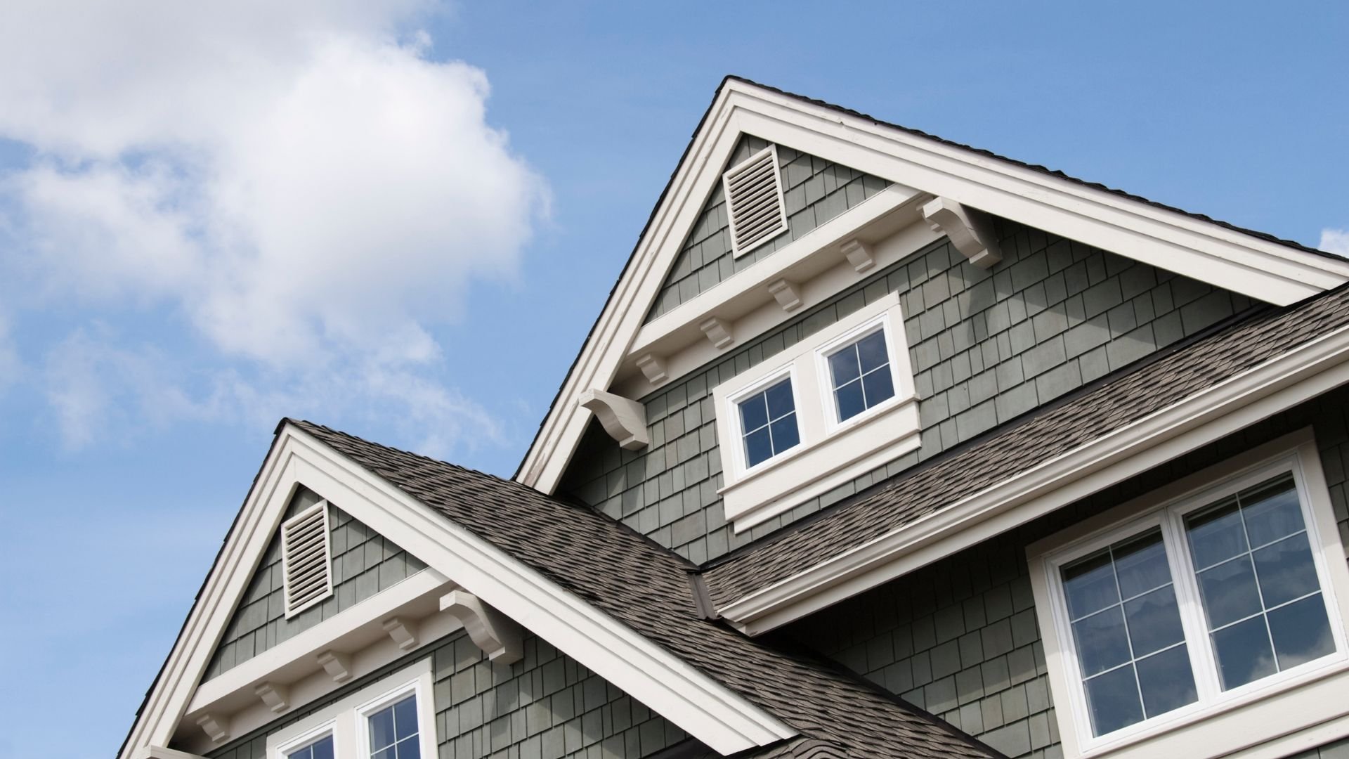 Gray shingled house with white trim and multiple windows against blue sky